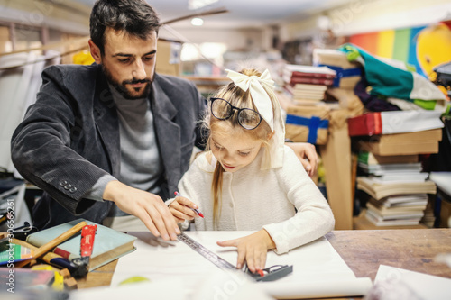Father sitting next to his beloved daughter and showing her how to make book bind. Gild sitting and using ruler and pen. Bookstore interior.