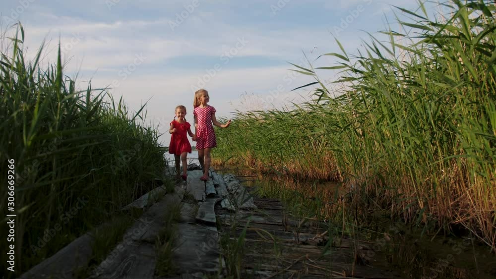 Two little cute girls red dress weared walk on old wooden pier through green grass near lake. Happy caucasian children walking by the lake and smiling. Childhood, freedom. Two little cute girls red dress weared walk on old wooden pier through green grass near lake. Happy caucasian children walking by the lake and smiling. Childhood, freedom.