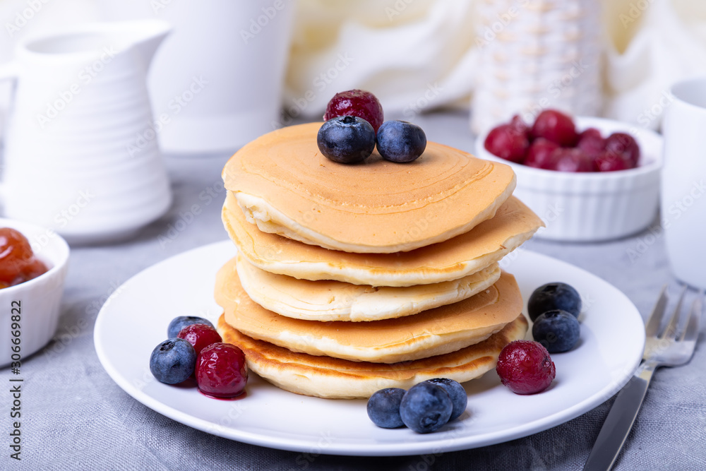 Pancakes with blueberries, cherries and mini apple jam. A cup of coffee and gravy boat in the background. Traditional american pancakes. Close-up. Pancakes with blueberries, cherries and mini apple jam. A cup of coffee and gravy boat in the background. Traditional american pancakes. Close-up.