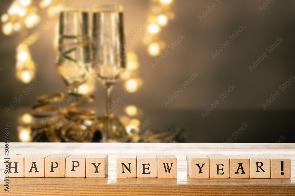 Happy New Year text made out of wooden blocks. Two glasses of champane with festive new year's eve decoration in the background. Copy space in the top right corner.
