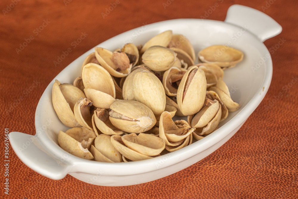 Lot of whole salted pistachio in small white ceramic bowl on cognac leather Lot of whole salted pistachio in small white ceramic bowl on cognac leather