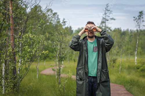 Canvas Print Young man in a dark green rain coat looks through binoculars with red lenses at