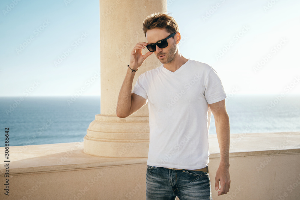 Young handsome guy is standing on a sea view background. Man is wearing white, empty basic t-shirt without logo. Horizontal mock-up with light flare. Young handsome guy is standing on a sea view background. Man is wearing white, empty basic t-shirt without logo. Horizontal mock-up with light flare.