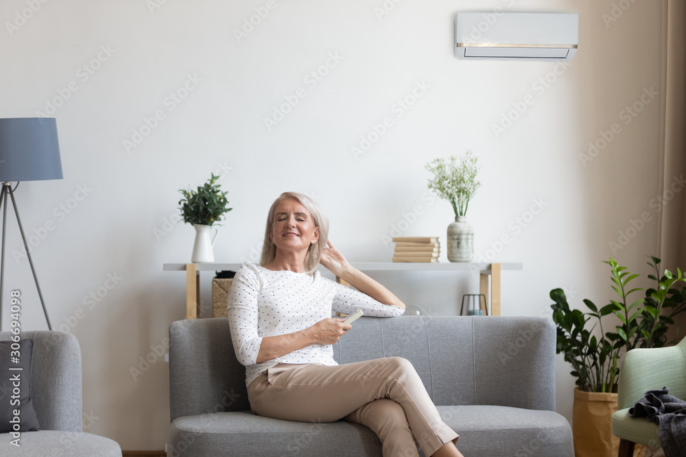 Aged woman sitting on couch holding remote control uses airconditioner Aged woman sitting on couch holding remote control uses airconditioner