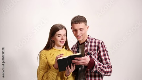 Young good looking smiling couple reads and turns pages of black notebook on the white background wall