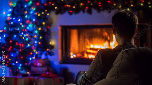 A young man admires the fire in the fireplace, which is decorated with garlands for Christmas. Back view