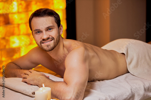 Portrait of young caucasian man lying on massage table at spa center with naked skin, massage on back and shoulders. Smile and look at camera, take pleasure from spa procedures