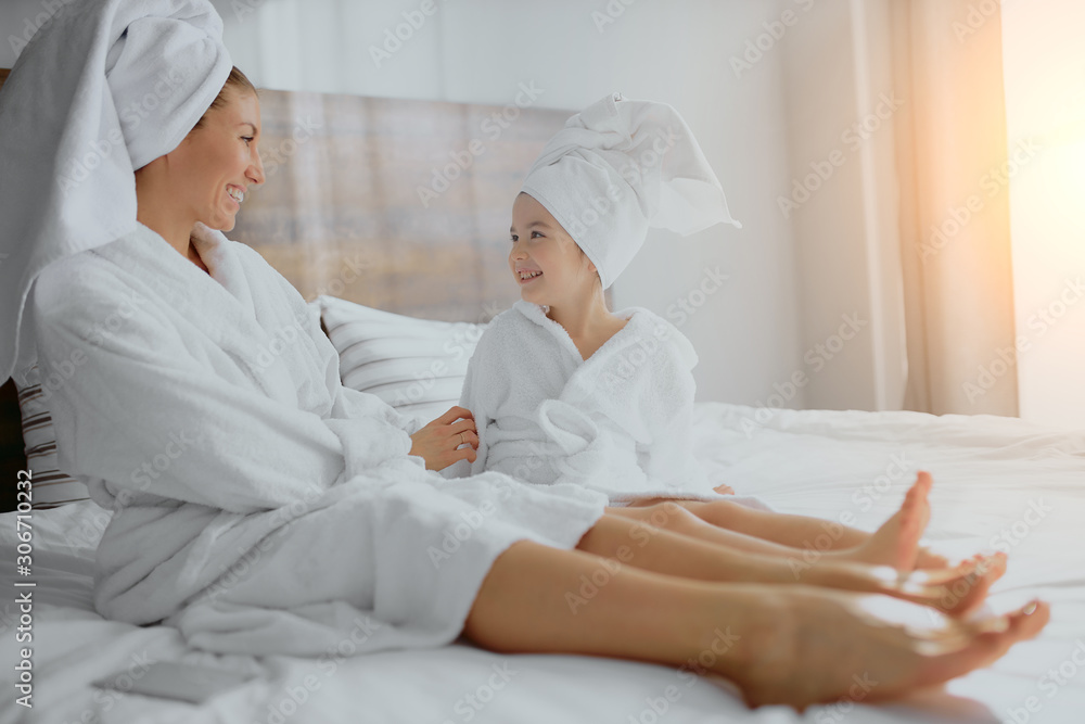 cute woman with little daughter sit together on bed in white towel and bathrobe after shower, look at each other cute woman with little daughter sit together on bed in white towel and bathrobe after shower, look at each other