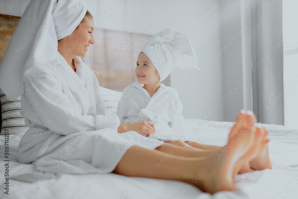 Fototapeta premium side view on lovely woman in white bathrobe and towel with cute daughter lying on bed in bright bedroom, little child look at mother