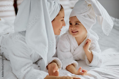 healthcare and family concept. caucasian young mother parent lie on bed with cute little girl kid child in bathrobe and towel, lovely look at each other