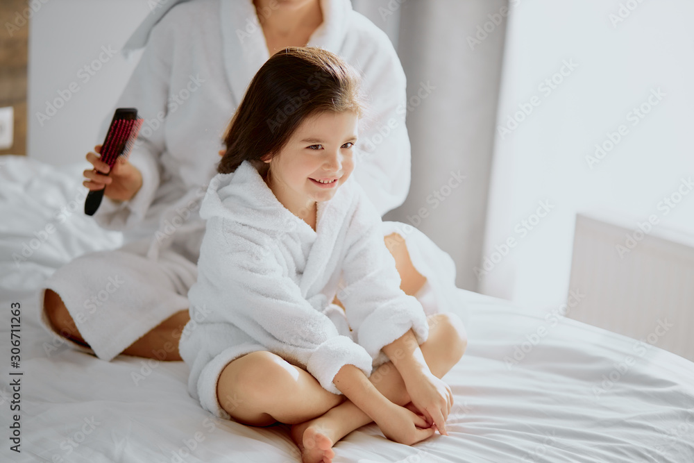 careful caucasian woman combing hair to awesome daughter dressed in bathrobe and towel after shower careful caucasian woman combing hair to awesome daughter dressed in bathrobe and towel after shower