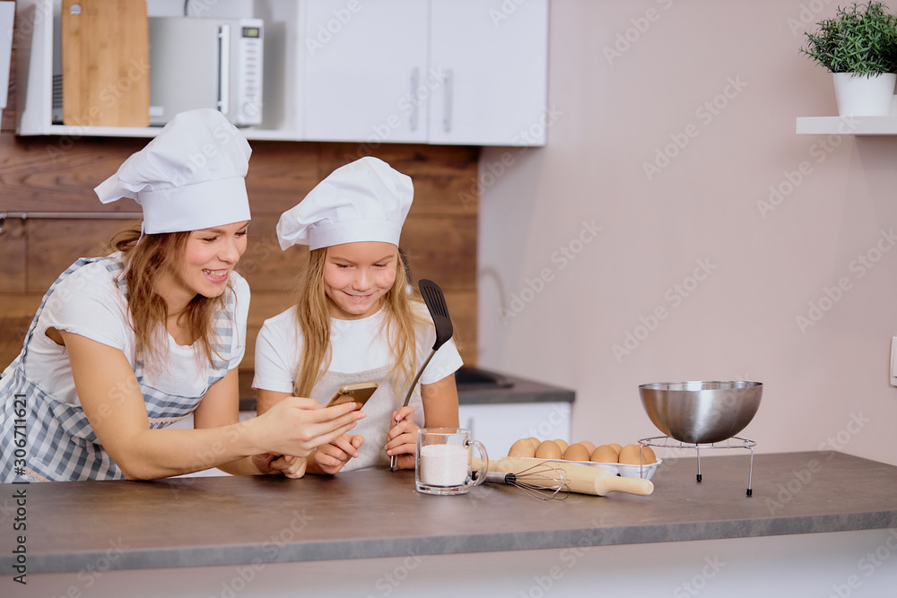 preparation for baking of mother and child, kid. cute daughter hold cooking spatula in hands and look at mobile phone, searching recipe of cake preparation for baking of mother and child, kid. cute daughter hold cooking spatula in hands and look at mobile phone, searching recipe of cake