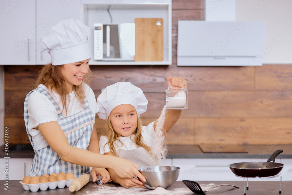 young woman mother baking with kid girl, daughter. little girl in apron and cap sprinkling flour in a bowl for making dough young woman mother baking with kid girl, daughter. little girl in apron and cap sprinkling flour in a bowl for making dough