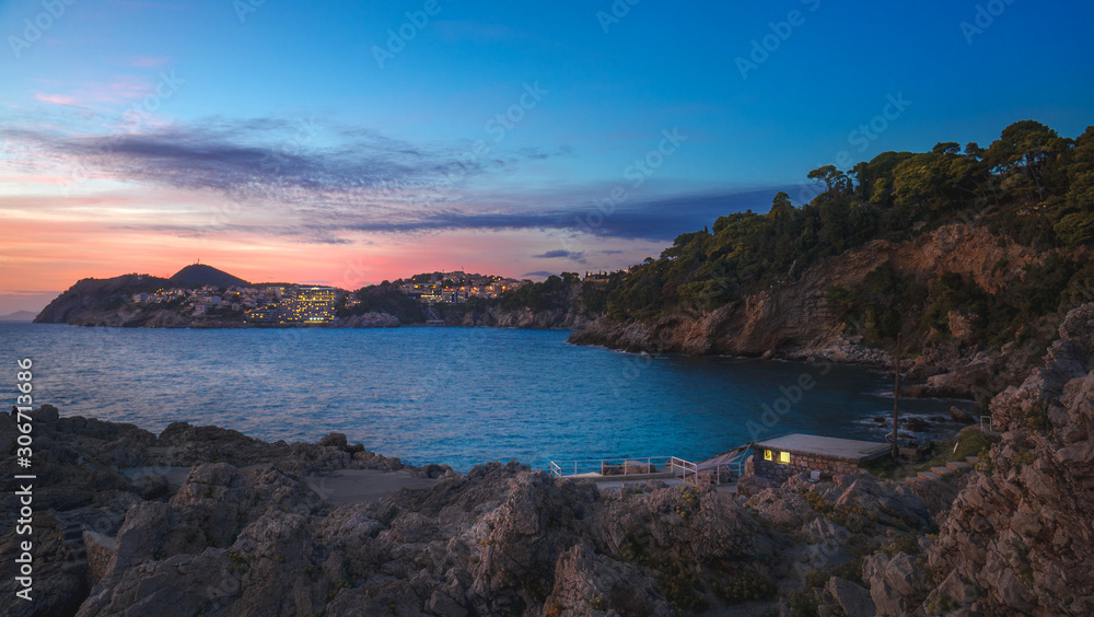 Rocky bay with lonely hut and night city lights on the background of scenic sunset sky