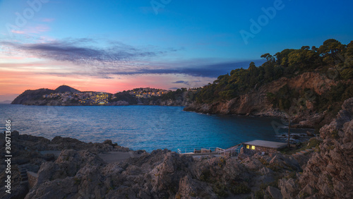 Rocky bay with lonely hut and night city lights on the background of scenic sunset sky