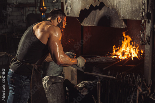 young caucasian strong man in black apron heating steel piece in furnace, powerful and strong muscles on hands, side view on caucasian man wearing gloves and eyeglasses for safety