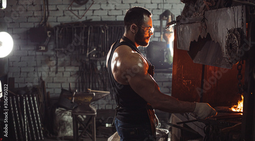 Portrait of a professional brutal caucasian blacksmith with strong muscles, side view on man opposite of fire