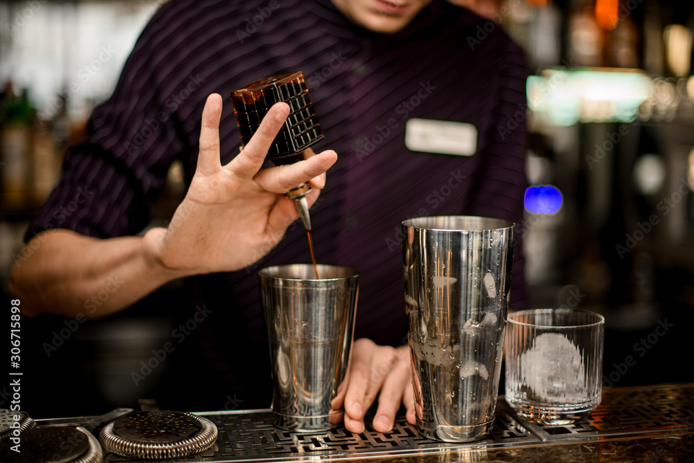 Professional bartender adding a brown bitter from a glass bottle to an alcoholic drink in a steel shaker