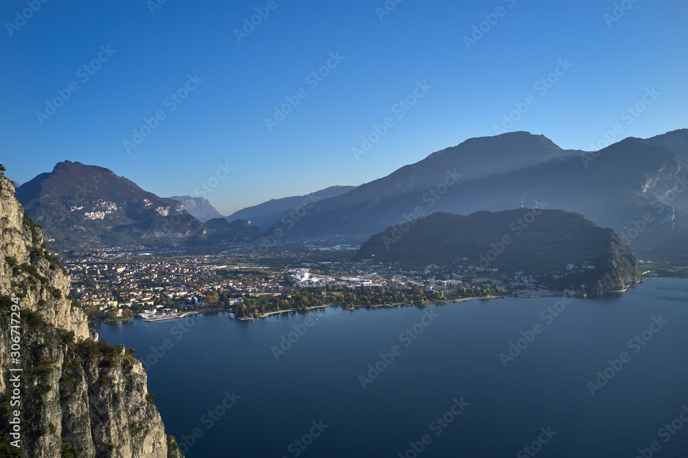 Steep cliff, view of the city of Riva del Garda, Italy. Panoramic view of Lake Garda in the foreground, the city is surrounded by rocks and alpine mountains. Autumn season.