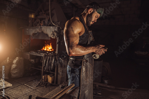 side view on forger man holding smartphone, having rest at work place. blacksmith shop owner, leisure time