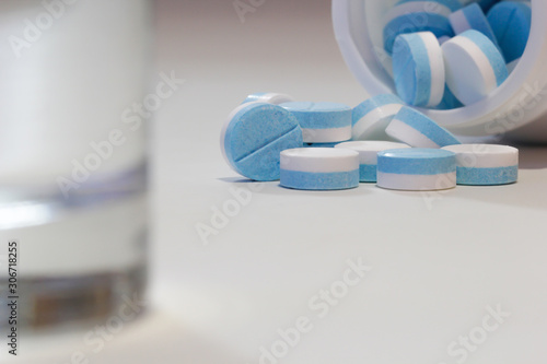 Blue and white pills and pill bottle with water glass on white table.