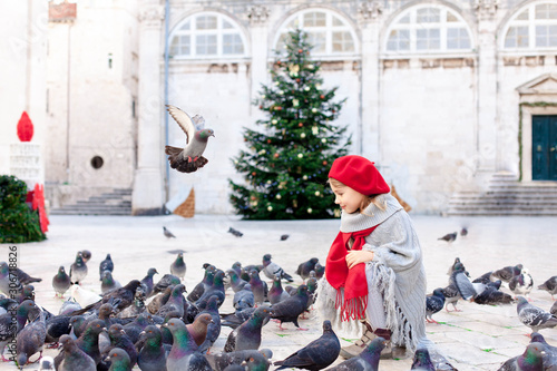 Christmas mood and holidays in Europe. Child girl feeds doves on square. Pigeons flying around cute kid. Romantic vintage atmosphere in old town Dubrovnik, Croatia. Authentic, ancient architecture.