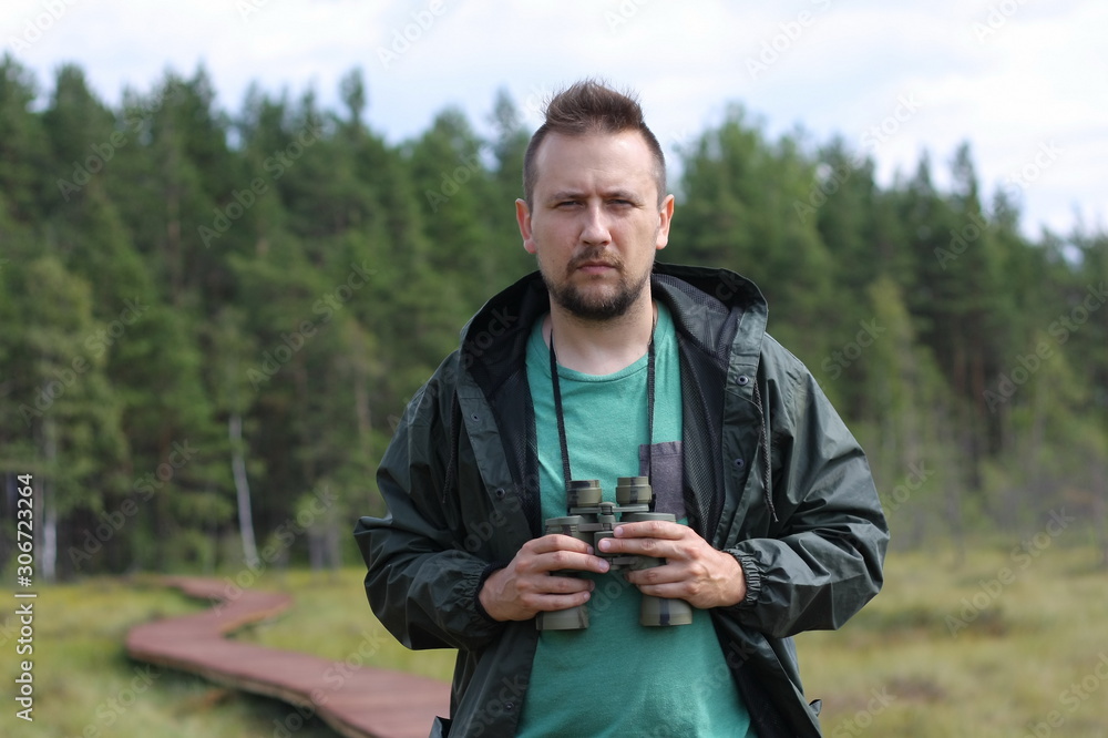 Portrait of a young man in a raincoat with binoculars in his hands against the background of a wooden path in the swamp Portrait of a young man in a raincoat with binoculars in his hands against the background of a wooden path in the swamp
