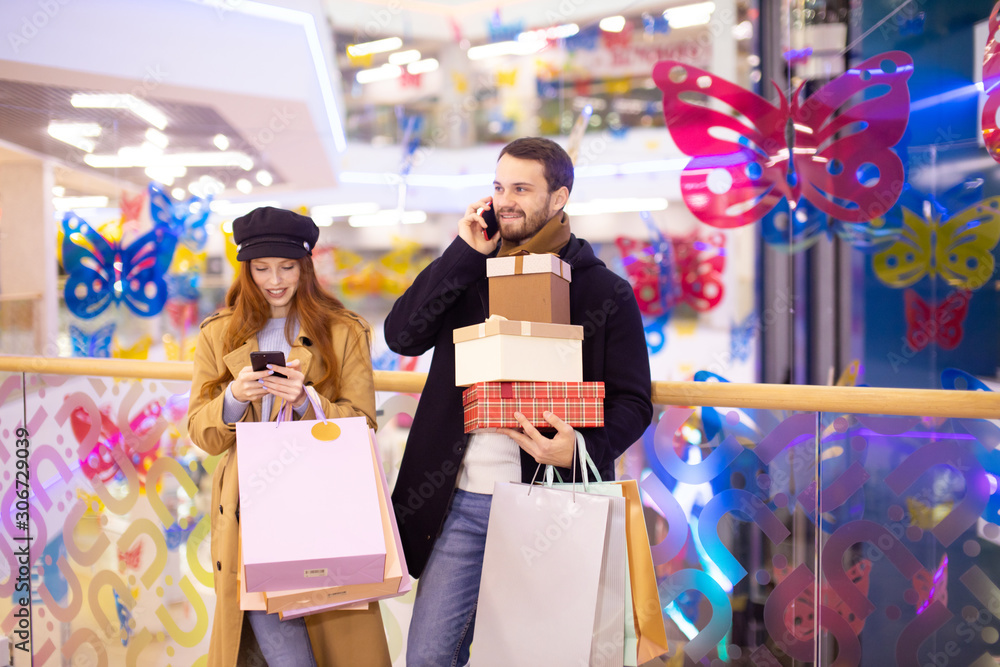 caucasian young couple with gifts and bags after shopping in shop center, redhaired woman and handsome bearded man together. man talking on phone, while his girlfriend look at screen of smartphone caucasian young couple with gifts and bags after shopping in shop center, redhaired woman and handsome bearded man together. man talking on phone, while his girlfriend look at screen of smartphone