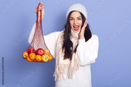 Excited young woman with fresh ripe mandarin, orange fruit isolated over blue background in studio, female wearing white winter cap and sweater, posing with opened mouth, keeps hand on cheek. photo