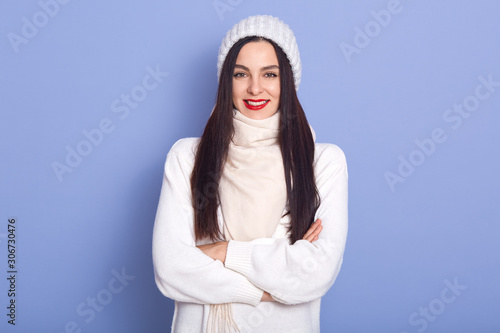 Good looking beautiful young woman with black straight hair standing isolated over lilac background in studio with crossed arms, having red lips, being fond of winter. Free time activities concept.