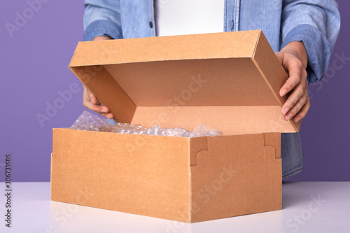 Faceless portrait of young man wearing denim shier and white shier opens brown carton box with present for his birthday, gift in case, male posing isoated over lilac studio backround. Holiday concept.