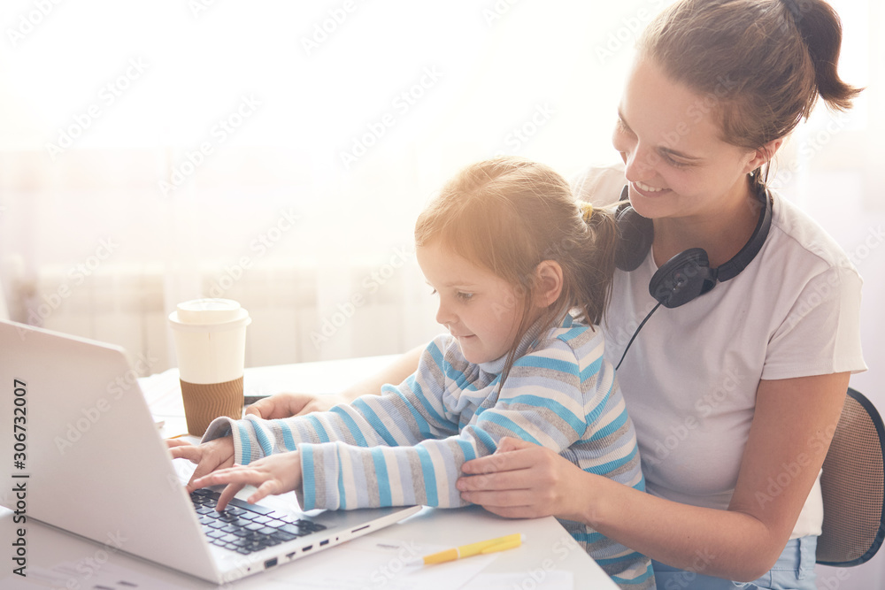 Happy family concept. Front view of adorable little girl sitting on mother  laps while watching cartoons online, mother and her child spending time  together, mom and kid wearing casual outfits. Stock-Foto |