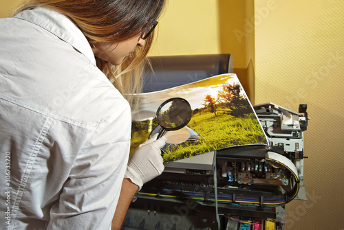 The girl in a white shirt prints a photo. Woman looking through photo with magnifier. The assistant checks the printed image. Nature, greenery and tent in the photo.