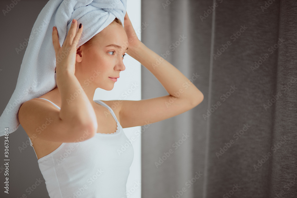 Woman with towel on head is relaxing in bedroom after shower, looking away with calm face, raising hands up, fixing white soft towel, background of gray cotton curtains Woman with towel on head is relaxing in bedroom after shower, looking away with calm face, raising hands up, fixing white soft towel, background of gray cotton curtains