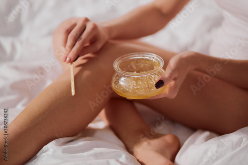 Gentle tender female hands holding glass jar with liquid sugar, putting wax on slender legs using wooden stick, close view, indoor hot, spa and epilation concept