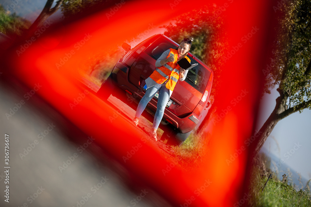 Naklejka premium Annoyed young woman in a road distress situation - setting up a warning triangle and calling for assistance after her car broke down in the middle of nowhere; transportation concept (car problem)