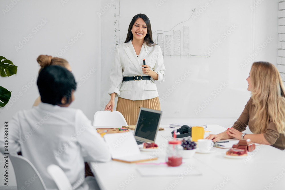 Fototapeta premium Beautiful successful business lady in white formal shirt holding felt pen smile while speaking with her colleagues listeners in office, flipchart behind her