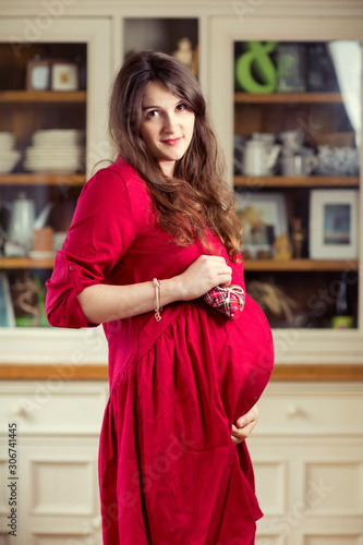 Pregnant women in red hat and red dress standing in front of rustic furniture . Christmas red dress