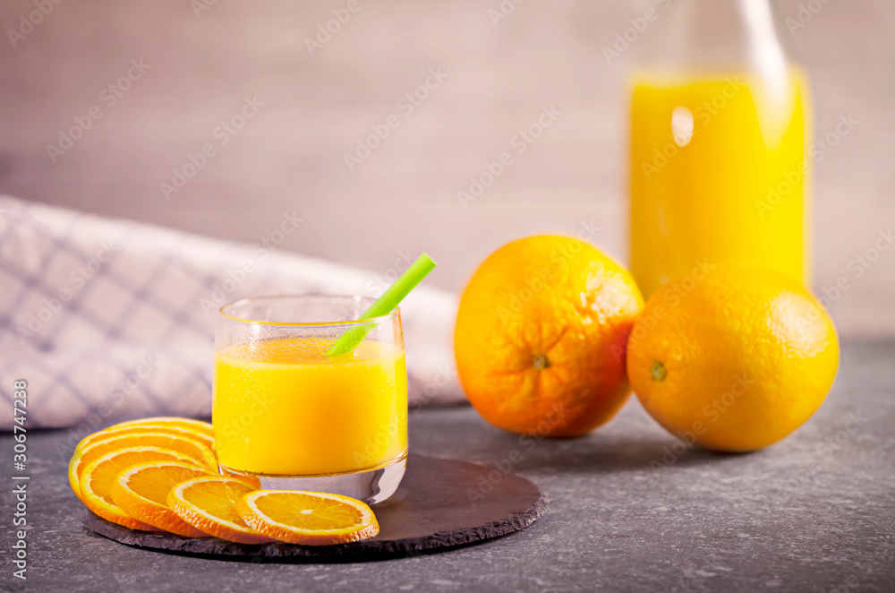 Fototapeta premium Delicious freshly squeezed orange juice in a small glass with fresh oranges, slices of orange and a bottle next to it on a rustic dark gray stone plate. Concept for healthy nutrition.