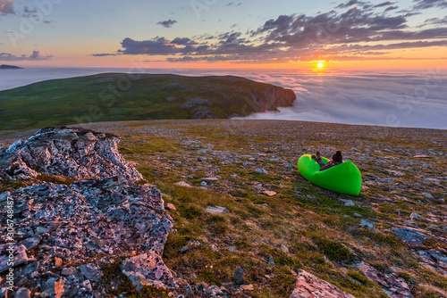 Woman in the air lounger is enjoying the midnight sun in the Northern cape of Soroya Island in Norwegian Finnmark. Fascinating sea of clouds is covering the Arctic Ocean surface at the background. photo