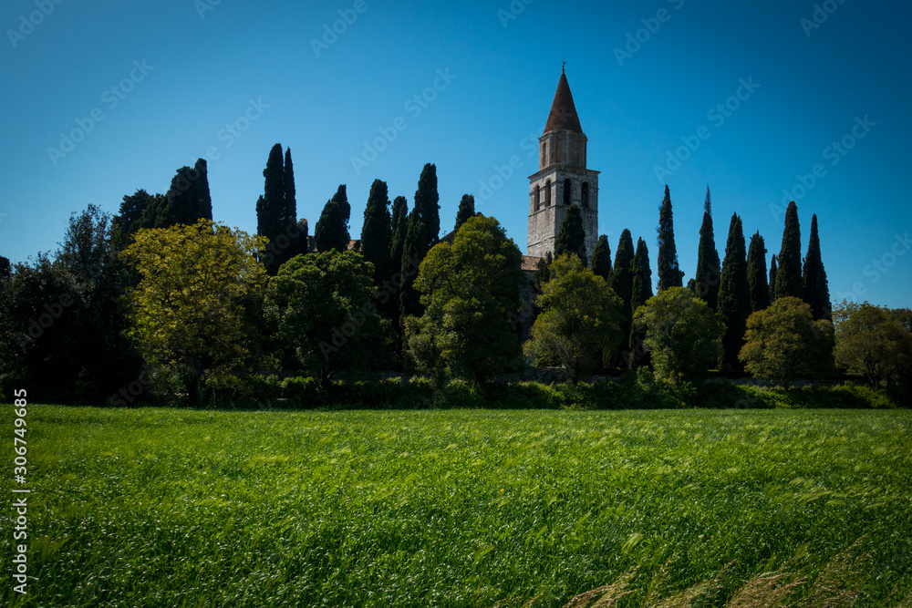 Tower of Basilica di Aquileia in Italy with meadows, trees, cypress and clear blue sky