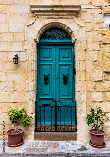 Ornate wooden green door in a stone entry in Cospicua, Malta. With glass window above, iron fence below and potted plants on both sides. Architectural theme.