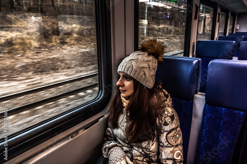Winter close-up portrait of beautiful young female looks out the train window. girl in winter clothes goes on vacation. She is wearing a gray jacket and knitted hat with a pompom.