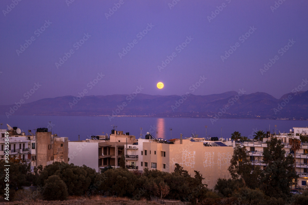 Purple dusk over the bay. Twilight in Hersonissos. Full Moon rises above the city. Cityscape by night. Buildings, Mediterranean Sea and mountains. Crete island. Greece.  Light reflections on the water