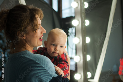 Attractive curly young mother in blue sweater hugging her toddler baby in red shirt in the room with Christmas decoration, Xmas tree and lights