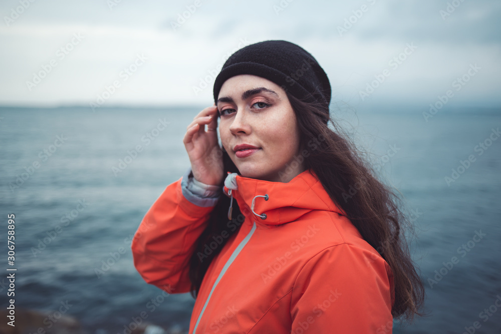 Portrait of a brunette woman with a mysterious look with long hair, wearing a hat and an orange jacket. In the background the sea and the horizon line