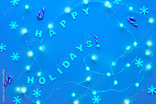 New Year or Christmas pattern flat lay top view background with text "Happy holidays". Paper snowflakes and garland of festive lights on blue background. Trendy monochrome toned background.