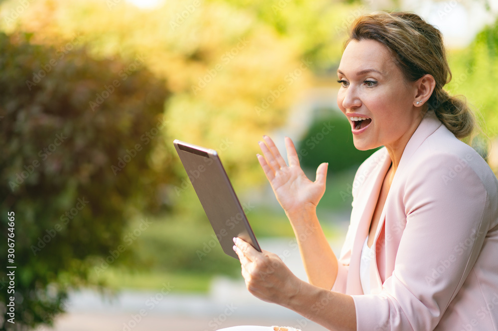 Excited businesswoman with a tablet in the park. Successful businesswoman is using tablet, outdoors. Adult woman communicating with a people uses tablet. Woman has a video call on gadget. Excited businesswoman with a tablet in the park. Successful businesswoman is using tablet, outdoors. Adult woman communicating with a people uses tablet. Woman has a video call on gadget.