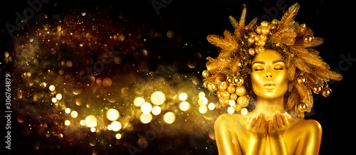 Christmas golden Woman. Winter girl pointing Hand, blowing blinking stars, Beautiful New Year, Christmas Tree Holiday Hairstyle and gold skin Makeup. Gift. Girl in decorated Xmas wreath. Beauty Model