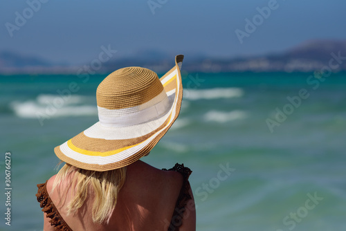 Young blond European woman in colorful sun hat standing against wonderful blue sea. She enjoying her rest at seacoast and looking towards sea.